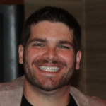 Corey Axelrod is a Caucasian man with brown hair. He is wearing a black shirt and tan sports coat. He is standing in front of a dark tan/brownish wall.