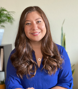 Alicia, an Asian woman with wavy brown hair, is wearing a blue blouse in front of a light grey background with plants.