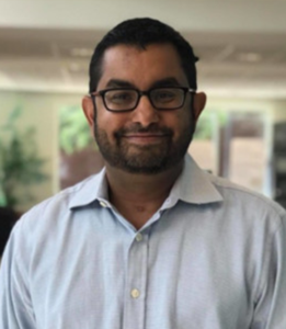 Alok Doshi, an Indian man with black hair, is wearing glasses and a light blue shirt. He is smiling in front of a blurred background.