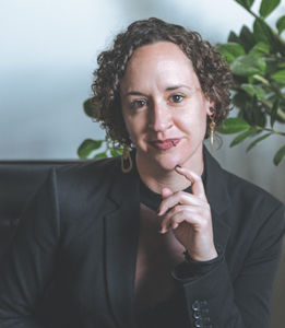 Bethany, a Caucasian woman with curly brown hair, is wearing a charcoal blazer and appears in front of a white background with a plant.