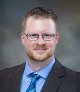 Dr. Christopher Moreland, a Caucasian man with reddish brown hair, is wearing blue glasses and a suit with a blue dress shirt and blue tie. He appears in front of a greyish background.