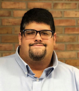 Jahan Farzam, a man of Middle Eastern descent with black hair and a goatee, is wearing glasses and a light blue shirt. He is pictured smiling in front of a brick wall.