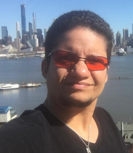 A Latino male looked at the camera smiling with city buildings, blue sky and blue water in the background. His hair is dark brown in a short crew cut style and chin hairs. He has orange glasses, silver rectangle pendant necklace and black shirt.