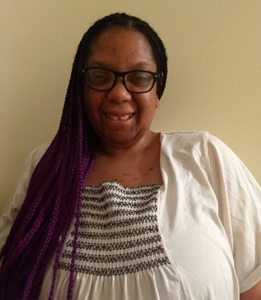 LeeAnne Valentine, a Black woman with purple braids, is wearing a white and grey striped blouse and a white cardigan. She appears in front of a white wall.