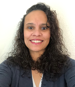 Natalie Burroughs, a biracial woman with curly black hair, is wearing a grey blouse. She is pictured smiling in front of a white wall.