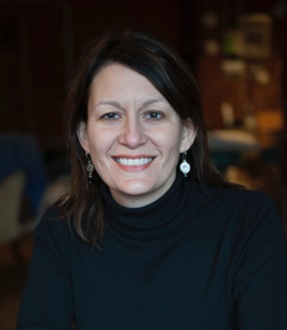 Trudy Suggs, a Caucasian woman with brown hair, is in front of a blurred background. She wears earrings and smiles at the camera. She is wearing a black sweater.