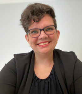 Debbie Mitre-Smith, a white woman with wavy brown hair, is in front of a white background. She wears glasses and smiles at the camera. She also wears a black top with a black cardigan.