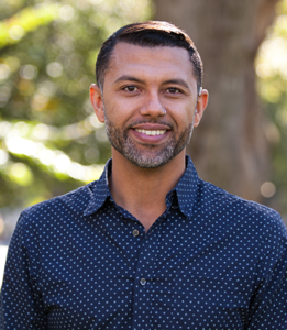 A brown man standing outside with hair combed to the side and short trimmed salt and pepper facial hair. He’s smiling at the camera wearing a navy blue button up with light polka dot prints. The background is blurred with trees, greeneries and people in the distance.