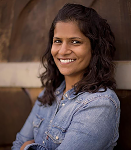 A woman with tan skin tone, dark brown and wavy hair, and blue jean shirt.