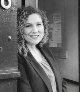 The image is a black-and-white photograph of Tiffany Ruffa with shoulder length curly hair smiling at the camera. She is standing in a doorway, dressed in a blazer and a top, with a professional and welcoming demeanor. The background shows part of the interior wall.