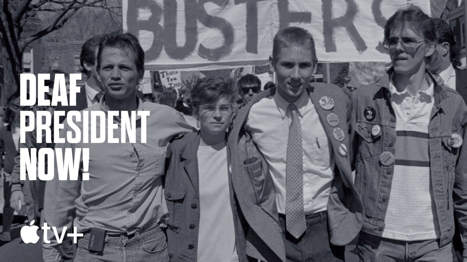 Black and white photo of the four leaders of the "Deaf President Now" protest, standing closely together outdoors with signs and buttons. "DEAF PRESIDENT NOW!" is overlaid in bold white text on the left, above the "tv+" logo. The individuals are dressed casually, suggesting a demonstration. A "BUSTERS" banner and another handwritten sign are visible behind them, with trees and a building in the background.