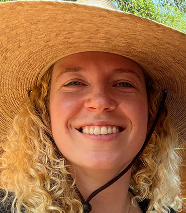 The image is a close-up, headshot-style photo of a woman with curly blonde hair, wearing a wide-brimmed straw hat. She is smiling broadly, showing her teeth, and looking directly at the camera. The lighting suggests an outdoor setting, possibly with sunlight.