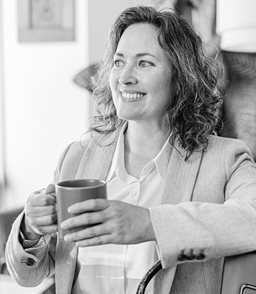 A black and white headshot of a woman with curly hair, wearing a light-colored blazer over a collared shirt. She is holding a mug and looking off to the side with a smile.