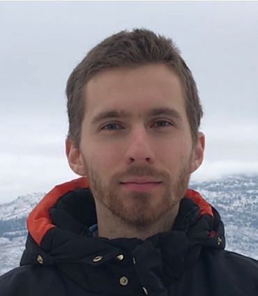 A headshot of a man with short brown hair and a beard, wearing a black and orange winter jacket. He is looking directly at the camera with a neutral expression. The background is a blurry, snowy landscape with mountains and trees.