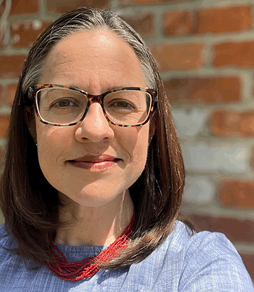 White woman with brown hair and glasses wearing a blue colored shirt and a red necklace shown from chest up again a brick background and sun shining on face.