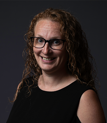 A close-up headshot of a woman with fair skin, curly red hair, and black-framed glasses. She is smiling directly at the viewer. She is wearing a black sleeveless top. The background is a solid dark gray.