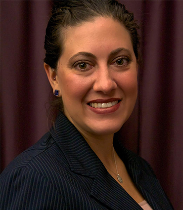 A headshot of Amanda David. She is smiling and looking directly at the camera. She has dark hair pulled back, dark eyes, and is wearing a dark pinstriped blazer over a lighter top, and a silver necklace with a circular pendant. There is a dark purple background behind her.