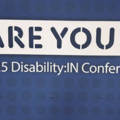 A man in a black polo shirt and dark pants stands to the left of a large sign that reads "ARE YOU IN?" The "IN?" part of the question is in green and blue. Below this, it reads "2025 Disability:IN Conference". The background is a dark blue with a subtle pattern of lighter blue dots. The man is smiling and wearing glasses.