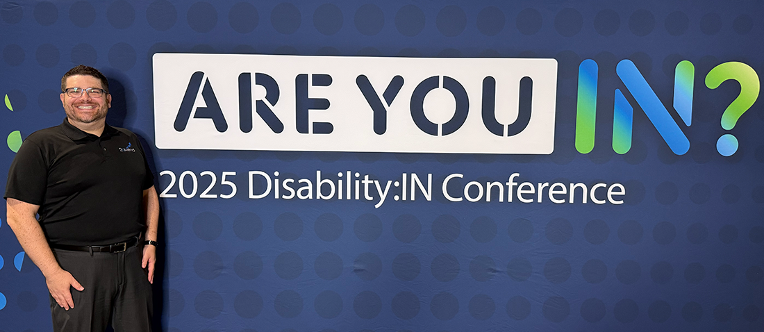 A man in a black polo shirt and dark pants stands to the left of a large sign that reads "ARE YOU IN?" The "IN?" part of the question is in green and blue. Below this, it reads "2025 Disability:IN Conference". The background is a dark blue with a subtle pattern of lighter blue dots. The man is smiling and wearing glasses.