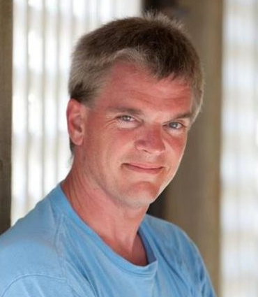 A close-up headshot of a man with fair skin and short, light brown hair. He is looking directly at the viewer with a slight smile. He appears to be middle-aged, with some faint lines around his eyes. He is wearing a light blue t-shirt.