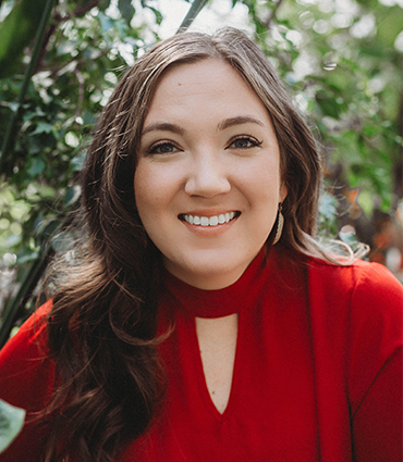 A headshot of Clara Baldwin, a light-skinned woman with long, dark brown hair, smiling directly at the camera. She is wearing a red top with a V-shaped cutout below the collar. Lush green foliage is blurred in the background.