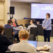 A "DOBE Roundtable" event is underway. Attendees are seated at tables in a conference room, engaged in discussions. Joyce Bender leads from the front while Corey Axelrod is addressing the group. In the background, the screen displays "DOBE ROUNDTABLE."