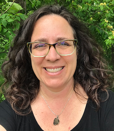 A headshot of Julie Stewart, a woman with curly brown hair and glasses, smiling against a backdrop of green foliage. She is wearing a black top and a necklace with a dark pendant.