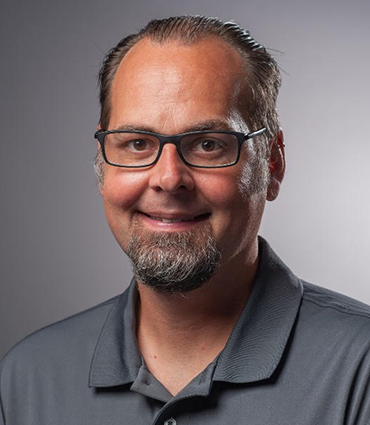 A headshot of Mark Davis, a man with short brown hair and a neatly trimmed goatee, is presented against a plain gray background. He is wearing glasses with dark frames and a gray collared shirt. He is smiling faintly and looking directly at the viewer.