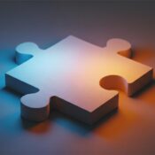 The image shows a single, three-dimensional puzzle piece rendered in a soft, matte material. It is illuminated by two distinct light sources, creating a gradient of color across its surface. One light source, coming from the left, casts a cool, blue-ish glow, while the other, from the right, emanates a warm, orange-yellow light. The interplay of these colors creates a subtle purple hue where they meet. The puzzle piece has a classic shape with two inward curves (holes) and two outward curves (knobs). The background is a simple, blurred surface that appears to be a continuation of the same material the puzzle piece is resting on, also affected by the dual lighting. The overall impression is one of simplicity and depth, highlighting the form and the effect of light.
