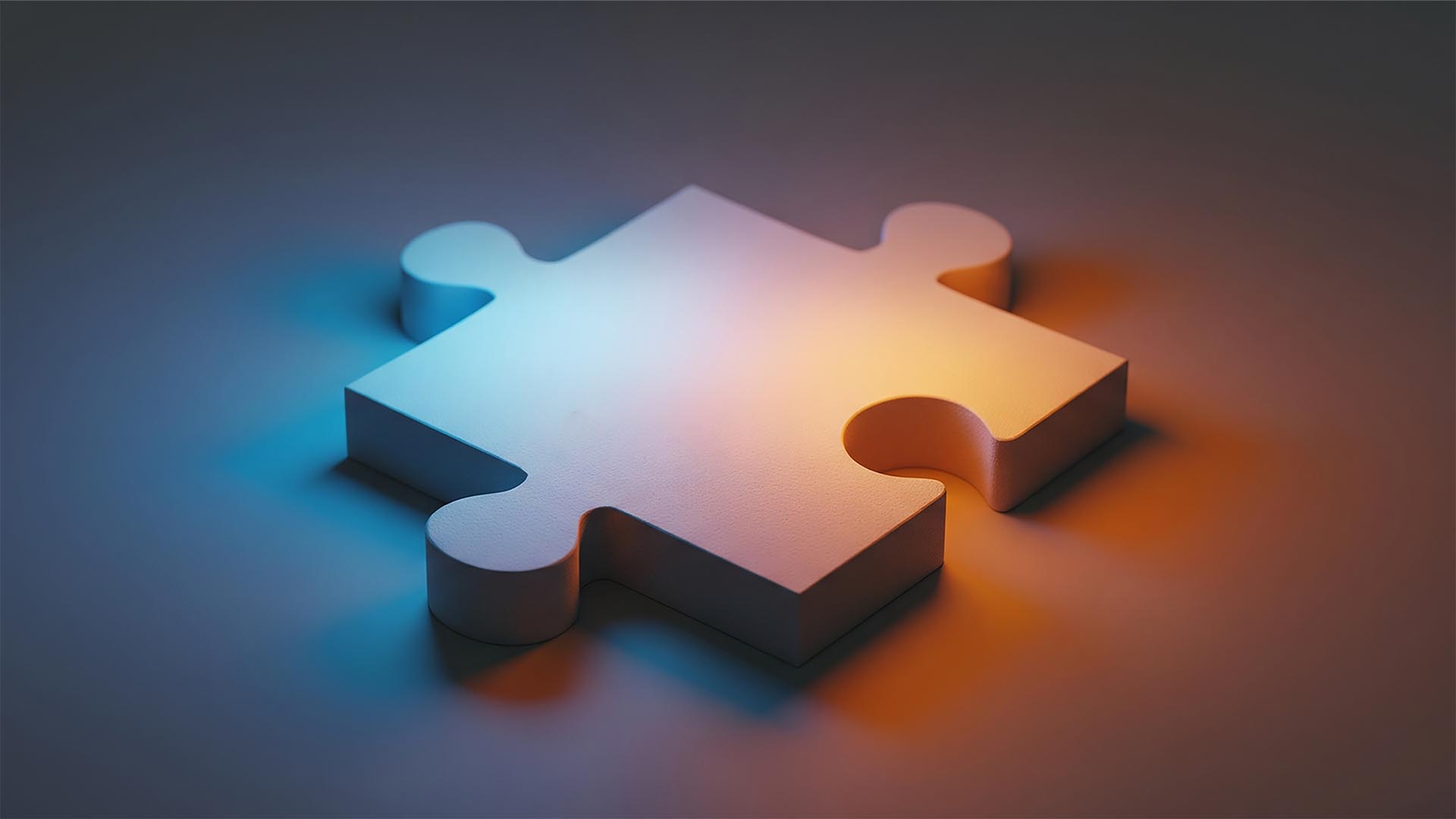 The image shows a single, three-dimensional puzzle piece rendered in a soft, matte material. It is illuminated by two distinct light sources, creating a gradient of color across its surface. One light source, coming from the left, casts a cool, blue-ish glow, while the other, from the right, emanates a warm, orange-yellow light. The interplay of these colors creates a subtle purple hue where they meet. The puzzle piece has a classic shape with two inward curves (holes) and two outward curves (knobs). The background is a simple, blurred surface that appears to be a continuation of the same material the puzzle piece is resting on, also affected by the dual lighting. The overall impression is one of simplicity and depth, highlighting the form and the effect of light.