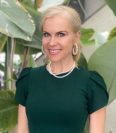 A headshot of Roberta Mather, a woman with blonde hair, fair skin, and blue eyes, smiling confidently at the viewer. She is wearing a dark green dress with puffed sleeves and a double-strand pearl necklace. Large, white, hoop earrings are visible. The background is softly blurred with green foliage.