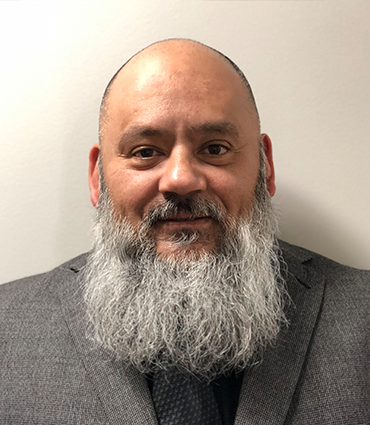 The image shows a headshot of a man, Tony Barraza.. He has a bald head and a prominent, full gray beard. He is wearing a dark grey suit jacket over a darker, possibly patterned, tie and a white shirt, suggesting a formal or business setting. The background is a plain, light-colored wall.