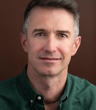 A professional headshot of a man with short gray and dark brown hair, fair skin, and blue eyes. He is wearing a forest green button-up shirt with the top button undone. He is facing the camera with a neutral, slight smile expression. The background is a solid medium-brown color.
