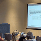 The image shows a presenter, Corey Axelrod, on a stage in a conference room, addressing an audience. A slide is projected onto a screen behind him, featuring two quotes in speech bubbles about the difficulty of getting medical accommodations and communicating at a doctor's office. The audience is seated and watching the presenter.