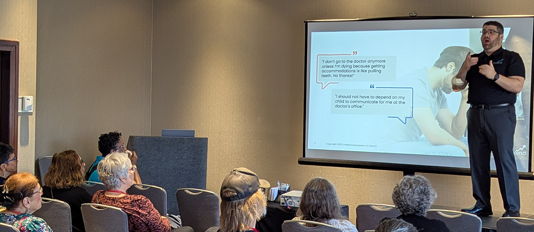 The image shows a presenter, Corey Axelrod, on a stage in a conference room, addressing an audience. A slide is projected onto a screen behind him, featuring two quotes in speech bubbles about the difficulty of getting medical accommodations and communicating at a doctor's office. The audience is seated and watching the presenter.