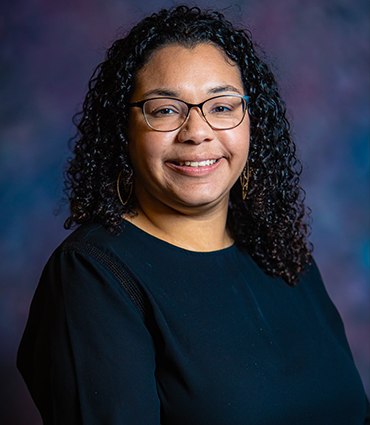Kellynette Gomez, a woman with dark curly hair and glasses, smiles at the camera. She's wearing a black top with a textured detail on the shoulder. The background is a gradient of purple and blue.