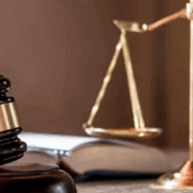A low-angle, eye-level shot shows a gavel and a scale of justice on a polished wooden surface. The setting appears to be a lawyer's desk or an old courtroom. The gavel lies in the foreground, angled to the left, with its head resting on a wooden base. The handle is short and rounded, and the head has a metal band. The scale of justice stands in the middle ground, on the right. Its trays are balanced and empty. A large, open book sits between the gavel and the scale, its pages blurred and unreadable. The background is a soft, blurry brown. The lighting is low and warm, casting a gentle glow on the objects.