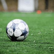 A soccer ball rests on a vibrant green artificial turf field, its white and dark blue geometric pattern standing out against the soft, blurry background. The image, shot from a low angle, captures the essence of the game with a focus on the well-worn ball, hinting at past matches and future play.