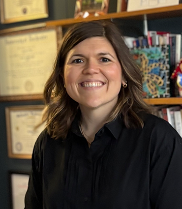 A professional headshot of a woman with shoulder-length brown hair smiling warmly at the camera. She is wearing a black collared shirt, positioned in front of a shelf displaying various framed certificates and books.