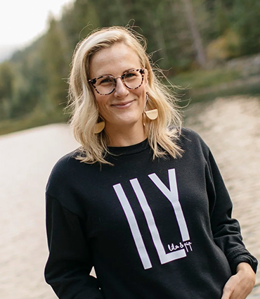 A smiling blonde woman with glasses wears a black sweatshirt with the letters "ILY" printed on the front, standing outdoors near a body of water.