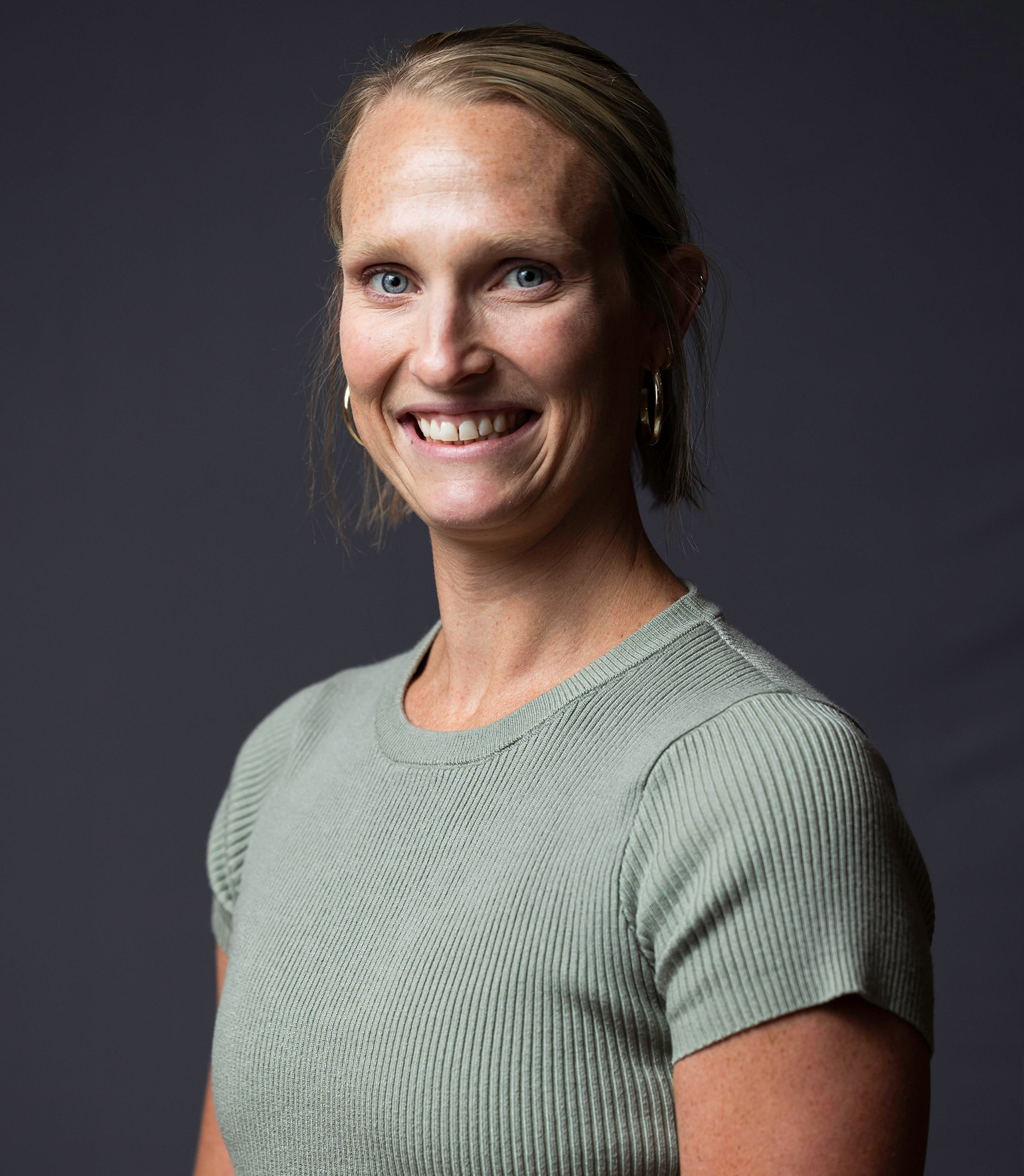 A professional headshot of Carly Weyers featuring her smiling directly at the camera against a simple, dark backdrop. She is wearing a light green ribbed top and gold hoop earrings.