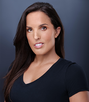 A professional headshot of Sarah Tubert, a woman with long, dark hair wearing a simple black V-neck top against a solid dark grey background. She is looking directly at the camera with a calm, confident expression, revealing a small hearing aid in her left ear.