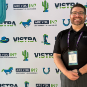 Corey Axelrod, Founder and CEO of 2axend, smiling while wearing a black polo shirt and conference lanyard in front of a branded step-and-repeat backdrop featuring the "ARE YOU IN? 2026 DISABILITY:IN CONFERENCE" logo, flanked by large decorative cactus props.