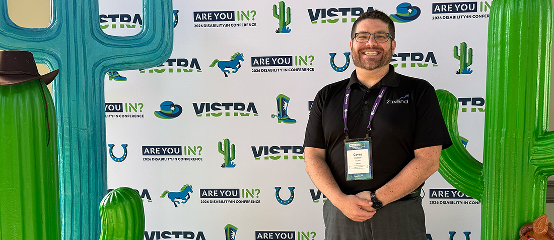 Corey Axelrod, Founder and CEO of 2axend, smiling while wearing a black polo shirt and conference lanyard in front of a branded step-and-repeat backdrop featuring the "ARE YOU IN? 2026 DISABILITY:IN CONFERENCE" logo, flanked by large decorative cactus props.