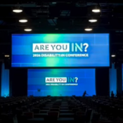 A wide shot of the main stage and auditorium at the 2026 Disability:IN Global Conference in Dallas, Texas. Three large display screens are mounted on the stage against a dark background, glowing with blue and green graphics that feature the text "ARE YOU IN? 2026 DISABILITY:IN CONFERENCE". Rows of empty conference tables and chairs face the stage in a large, darkened ballroom.