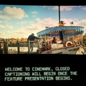 A movie screen at a Cinemark theater shows a scenic movie still of a boat docked near a pier beneath a partly cloudy sky. Below the main screen, a specialized electronic captioning display reads, "WELCOME TO CINEMARK, CLOSED CAPTIONING WILL BEGIN ONCE THE FEATURE PRESENTATION BEGINS."