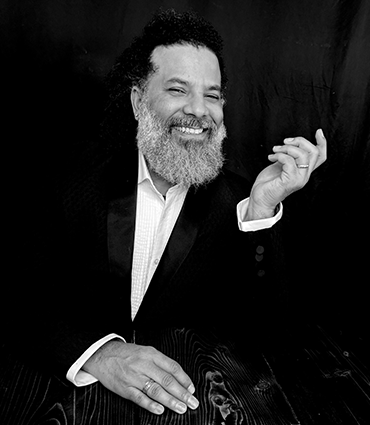 A black-and-white headshot of Jeremy L. Brunson smiling warmly while wearing a dark suit jacket and a white collared shirt. He has a full beard and mustache, and his left hand is raised near his shoulder with fingers slightly curled.