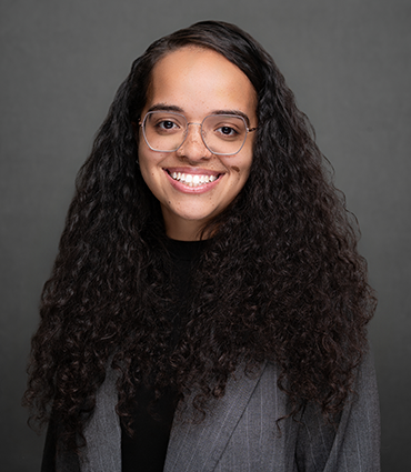 Naima Johnson smiles warmly while facing the camera against a textured grey background. She wears light-colored wire-rimmed glasses, a black shirt, and a grey blazer over her long, dark curly hair.