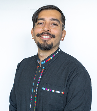 Headshot of Topher Ávila smiling warmly against a white background. He wears a black shirt featuring colorful, patterned trim along the collar and front button placket.