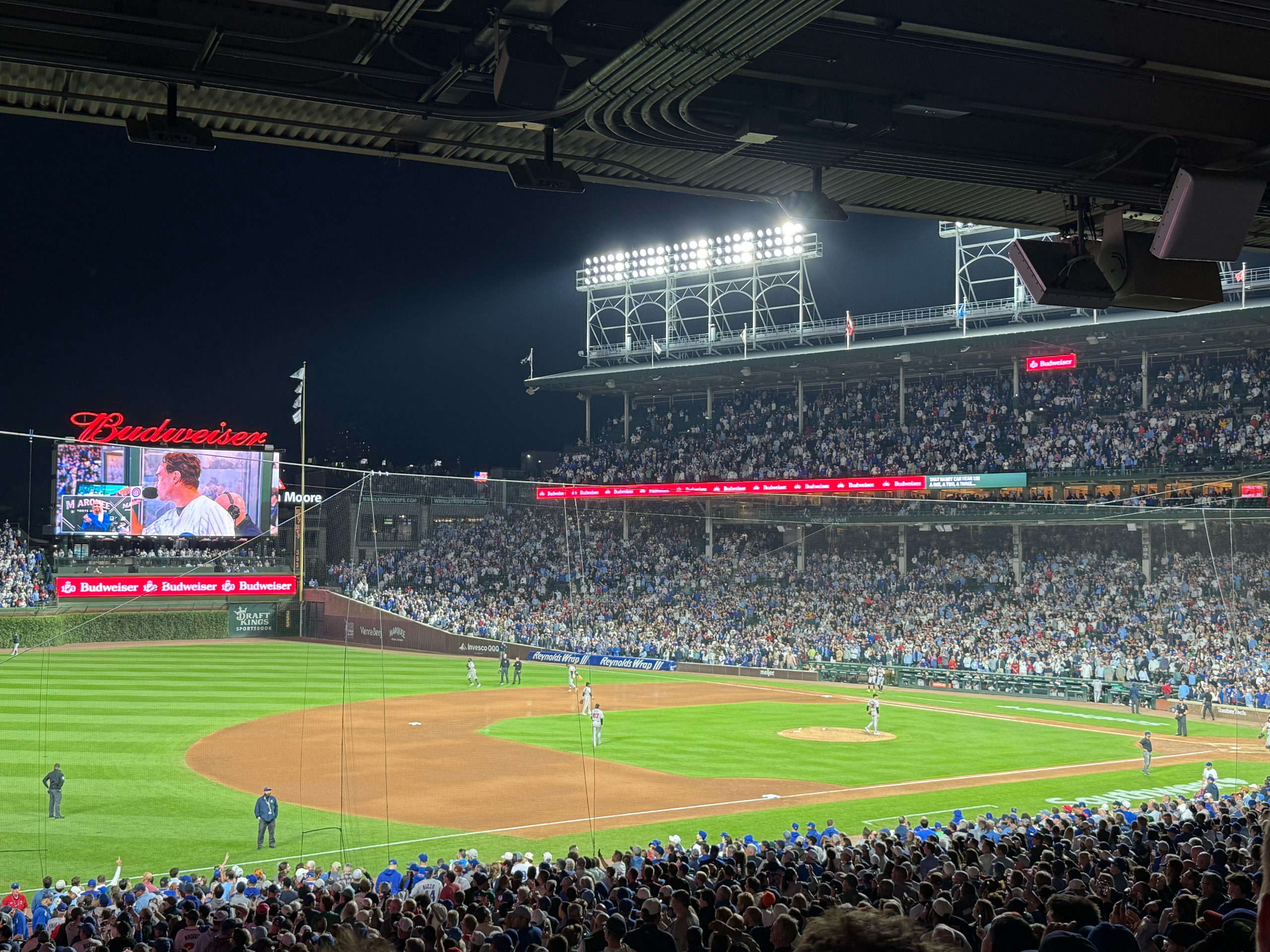 A view of a night baseball game from lower-deck seats near the third-base line, looking across the illuminated infield toward the pitcher's mound and right field. Bright stadium light towers tower over the grandstands, and the right-field Jumbotron displays a live broadcast feed above the outfield wall as spectators fill the surrounding seats.